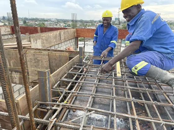Two builders in hard hats tying steel reinforcement for a floor slab, with the city spread out behind them.