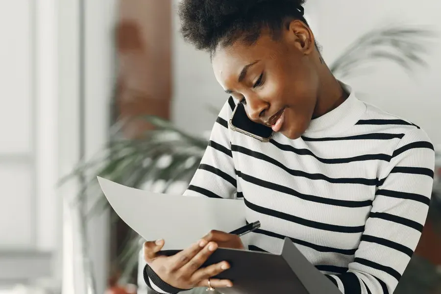 A woman in a striped top takes notes on a paper form while on a phone call.