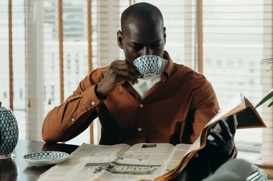 A man in a rust-coloured shirt sips from a patterned cup while reading a newspaper at a polished table.