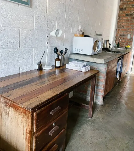 A studio kitchenette: a worn timber desk with a lamp beside a concrete counter holding a microwave, kettle and sink, against a white block wall.