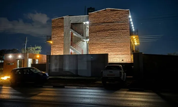 The OSK Kalingalinga building at night, its brick walls and open stairways lit, with two cars parked on the road in front.