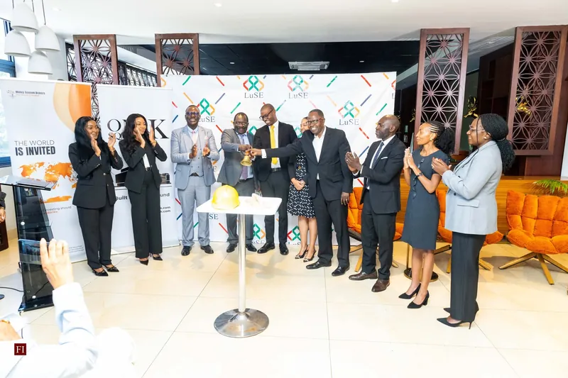 Representatives of OSK, Money Acumen and the LuSE ring a bell beside a gold hard hat, in front of a LuSE backdrop, as colleagues applaud.
