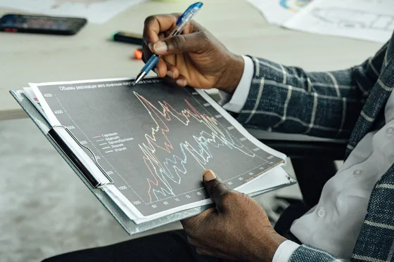 A hand points a pen at a printed line chart on a clipboard.