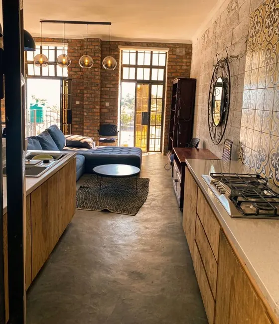 A long kitchen with timber cabinets and a gas hob leading to a lounge with a blue sofa and tall steel-framed windows.