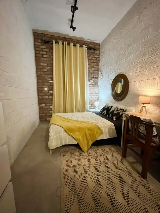 A narrow furnished studio with an exposed brick wall, mustard curtains, a round mirror, a desk and a patterned rug.