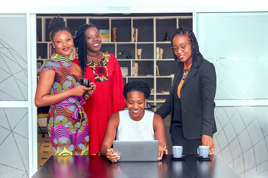 Four smiling women in a bright office, three standing and one seated at a laptop, with coffee cups on the table.